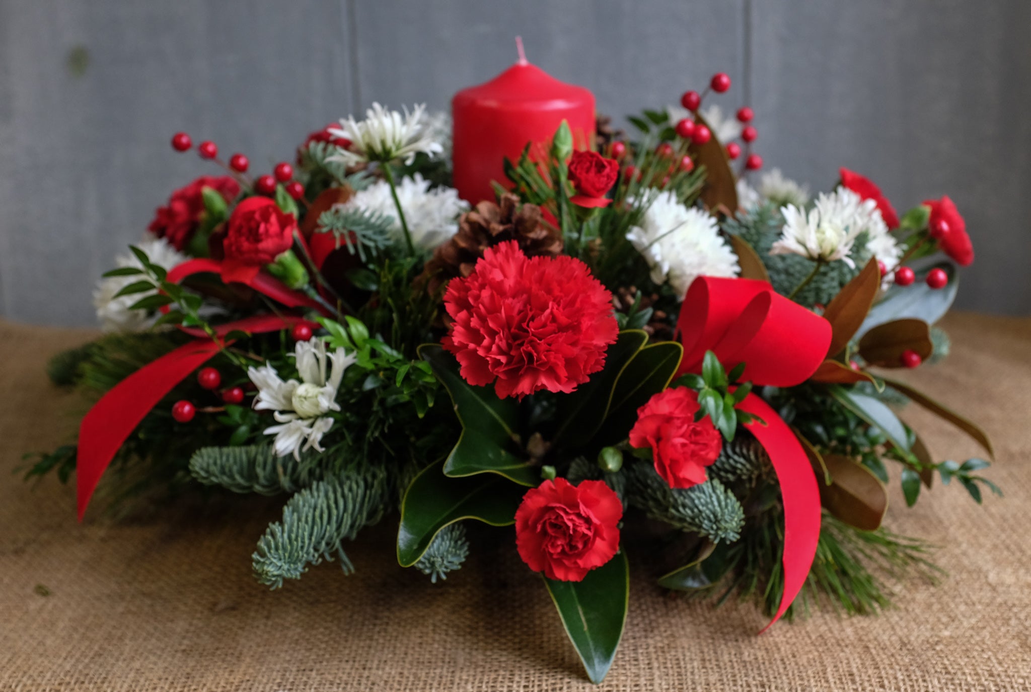 Floral centerpiece with carnations, mums, red bows, magnolia leaves, pine cones, and red pillar candle by Michler's Florist