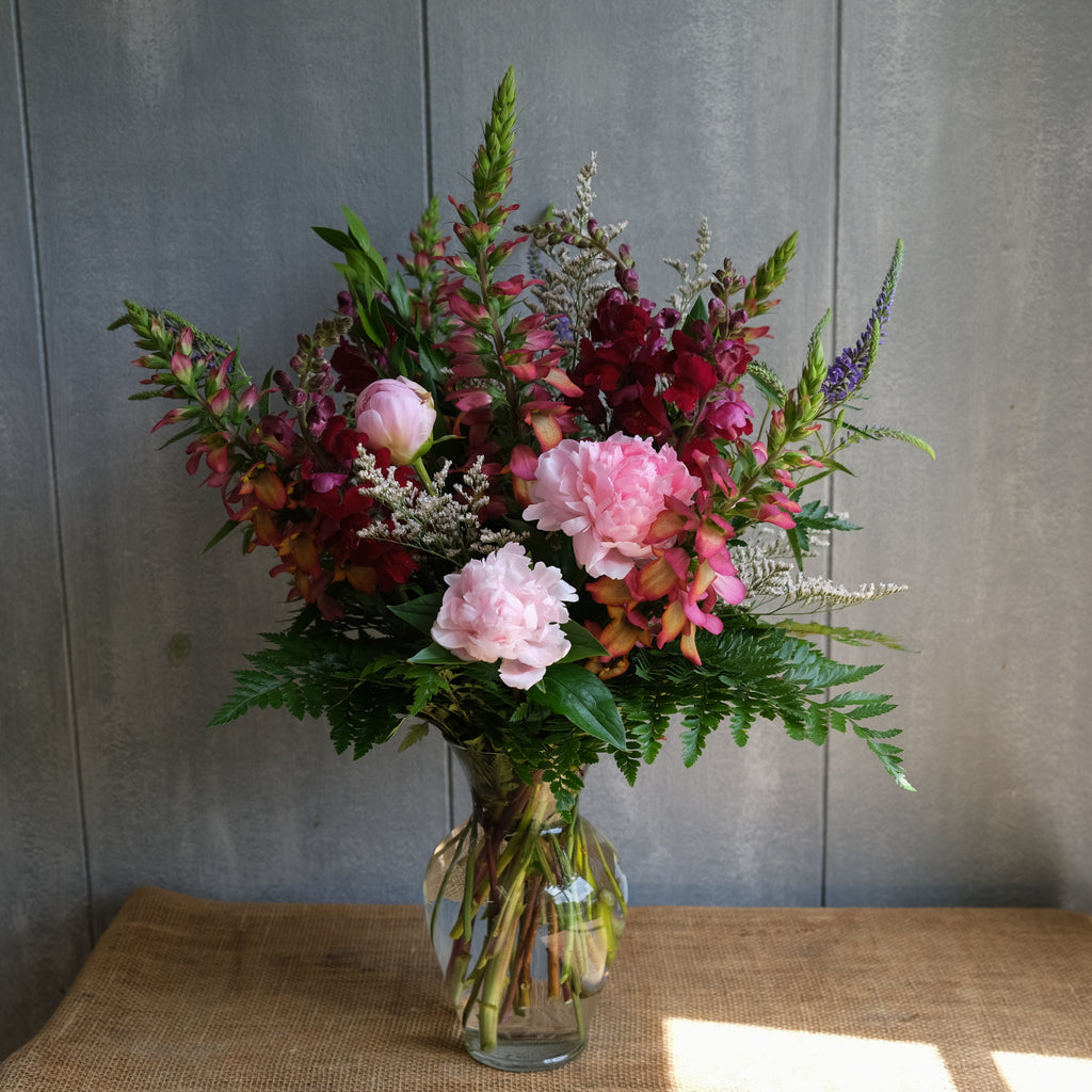 flora arrangment in a tall design with peonies, digiplex, veronica, snapdragons, all in pink, maroon, and purple colors