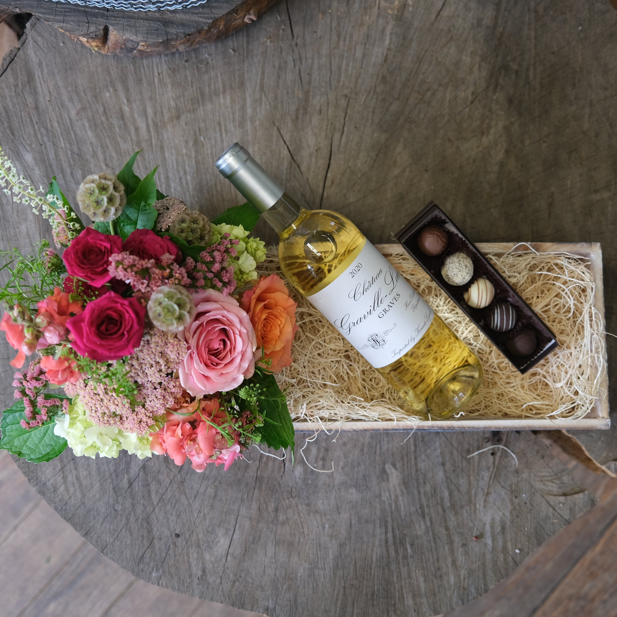 Oblong wooden crate featuring a bottle of white wine, a box of five truffles, and a warm, compact flower arrangement.